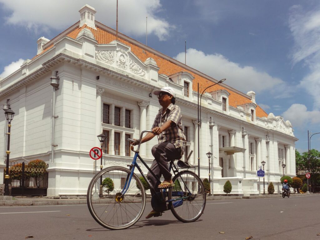 A man riding a bicycle past a historic colonial building in Surabaya, Indonesia, under a sunny sky.